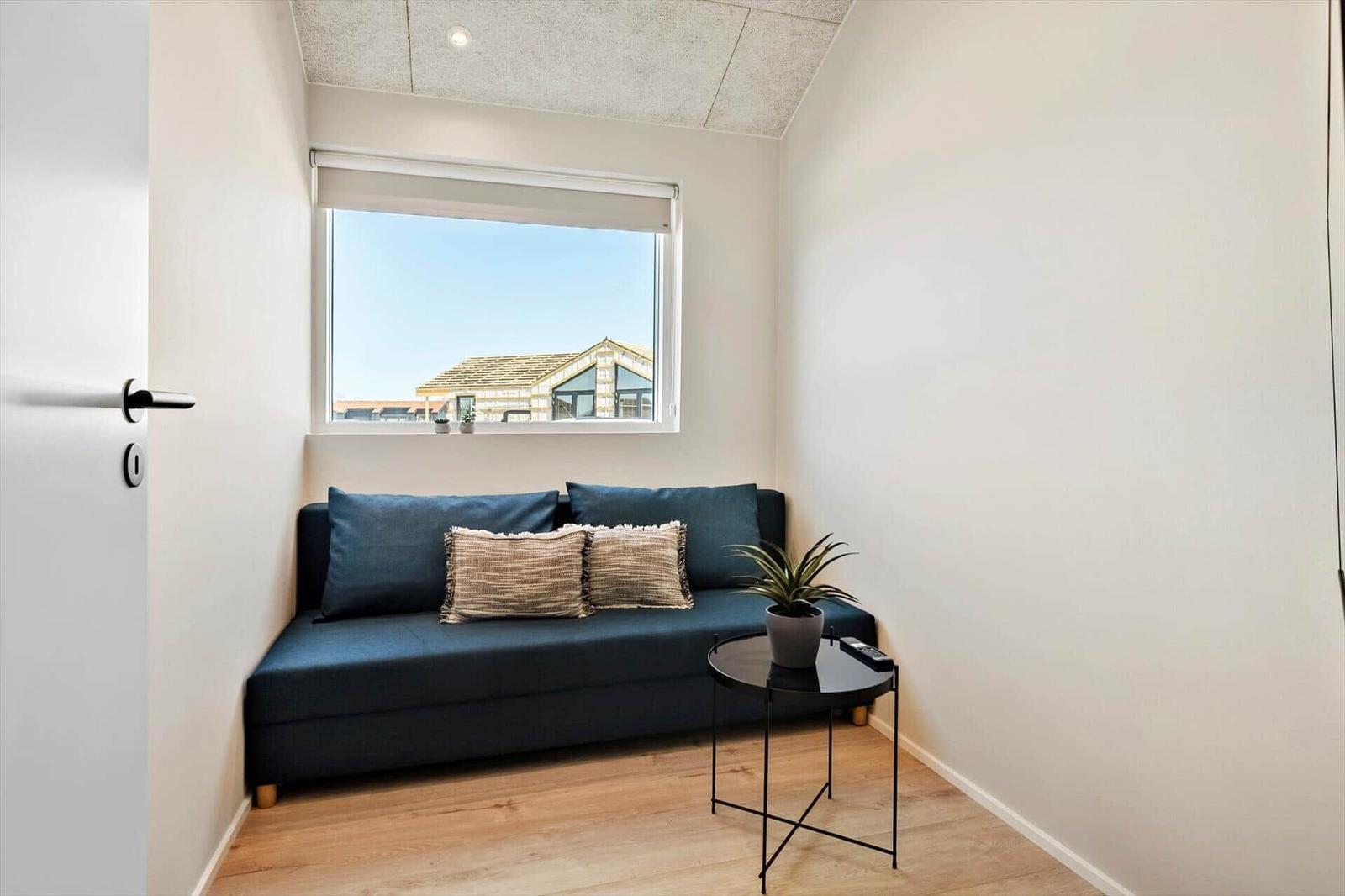A room with blue sofa, small table and plant. Window shows roof and sky.