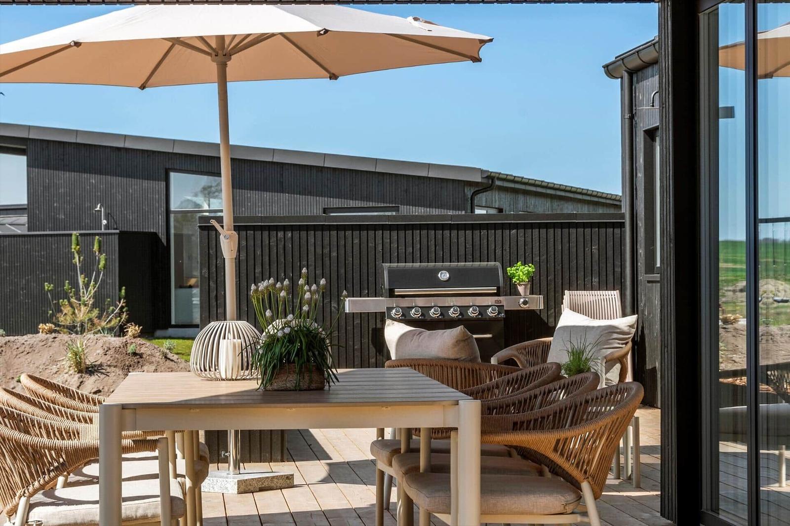 Terrace with table, chairs, umbrella, and grill. Background: black wooden fence and building.