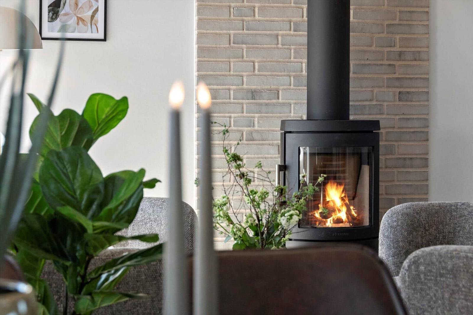 A wood-burning stove with a fire is set against a brick wall. Two candles and a plant are in the foreground.