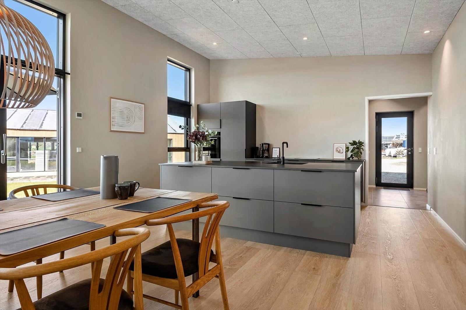 Modern kitchen with dining area and large windows. Wooden floor and gray cabinets.