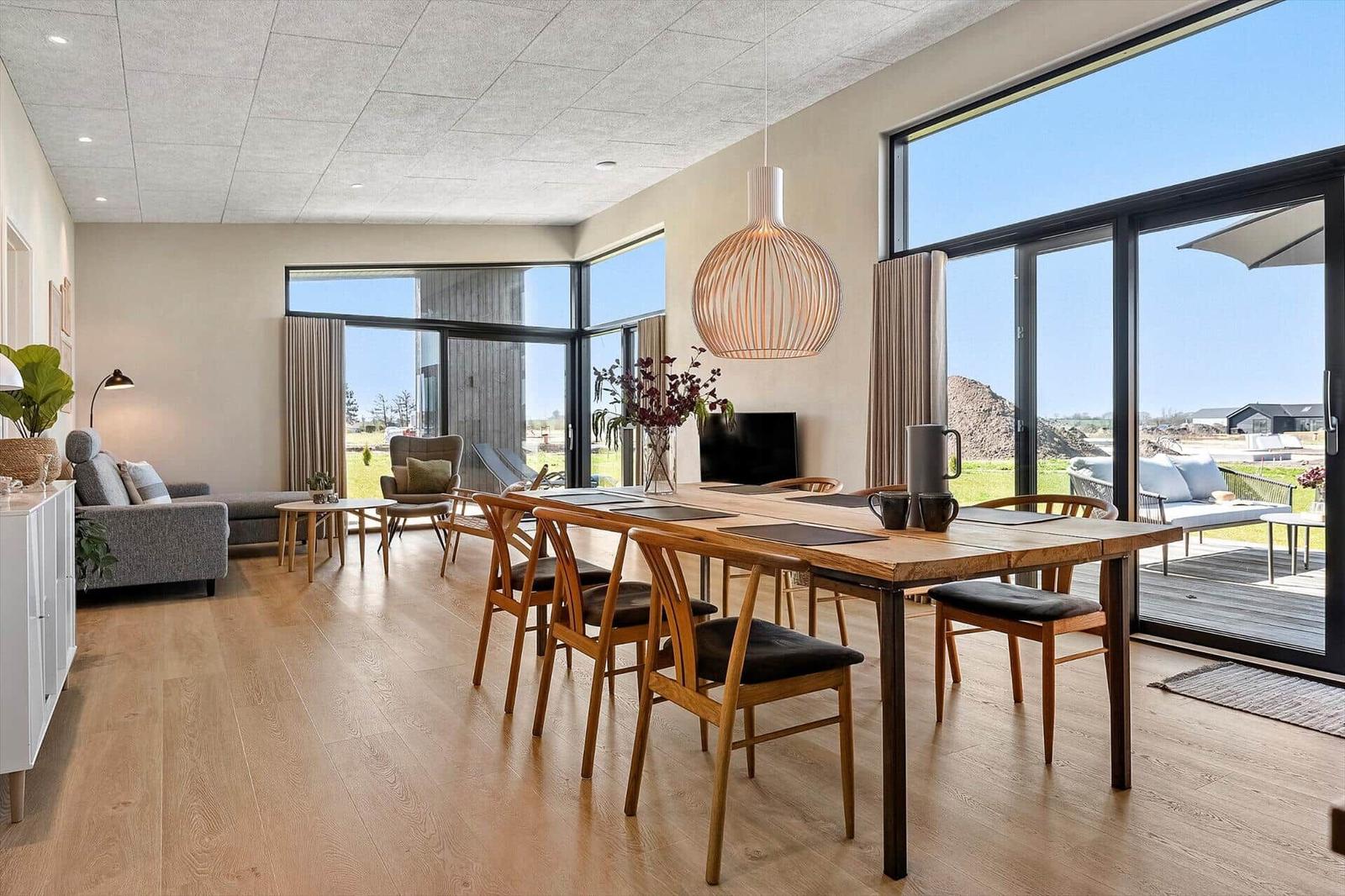 Dining area with wooden table and chairs, large windows, and terrace overlooking green space.