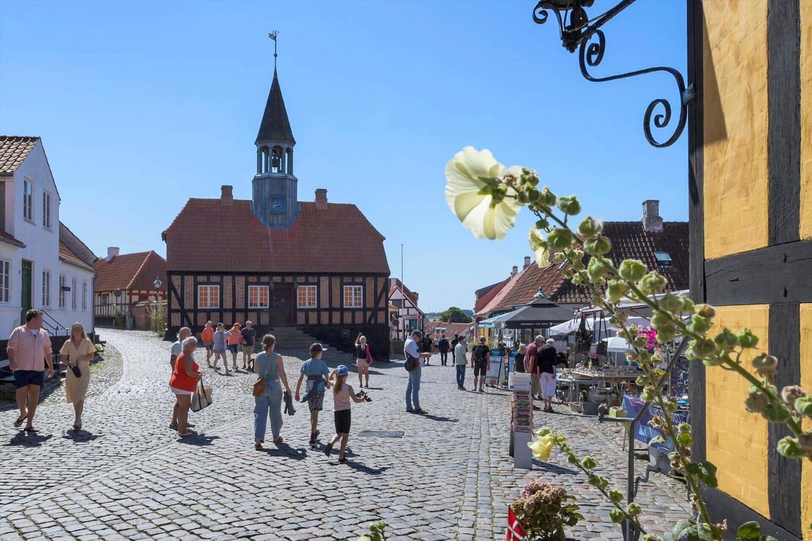 Cobbled street with historic town hall and market stalls. People stroll on stone pavement.