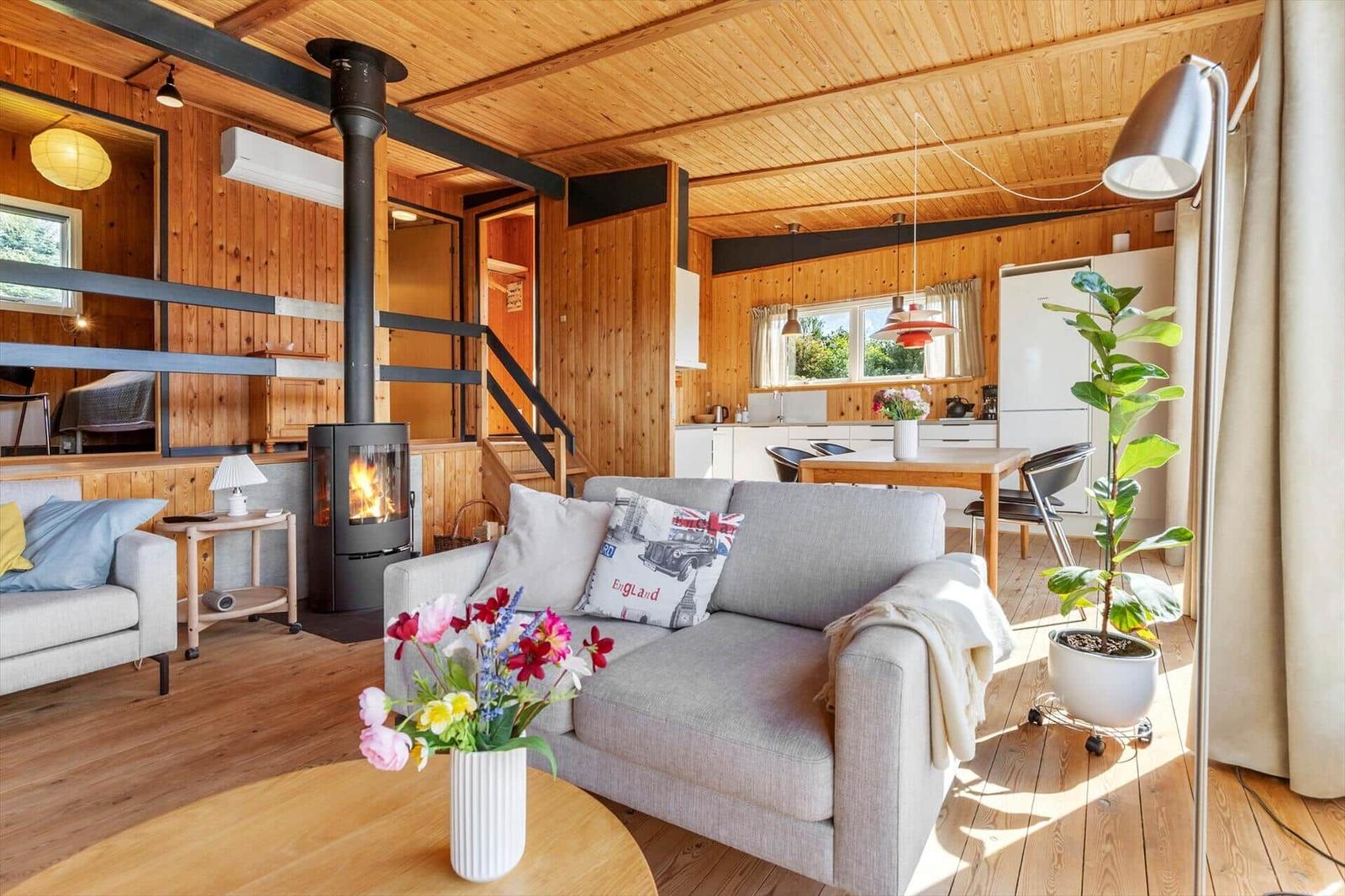 Living room with fireplace, sofa, and kitchen area. Wooden floor and ceiling.