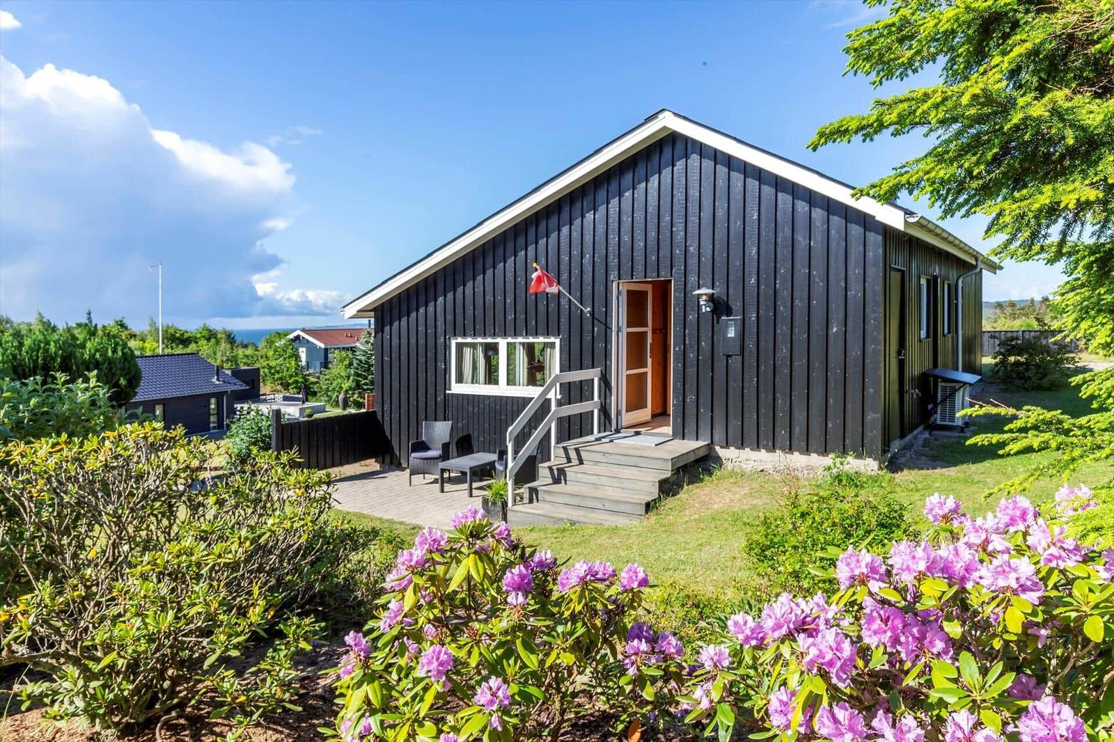 Black wooden house with terrace, blue sky, and blooming rhododendrons.