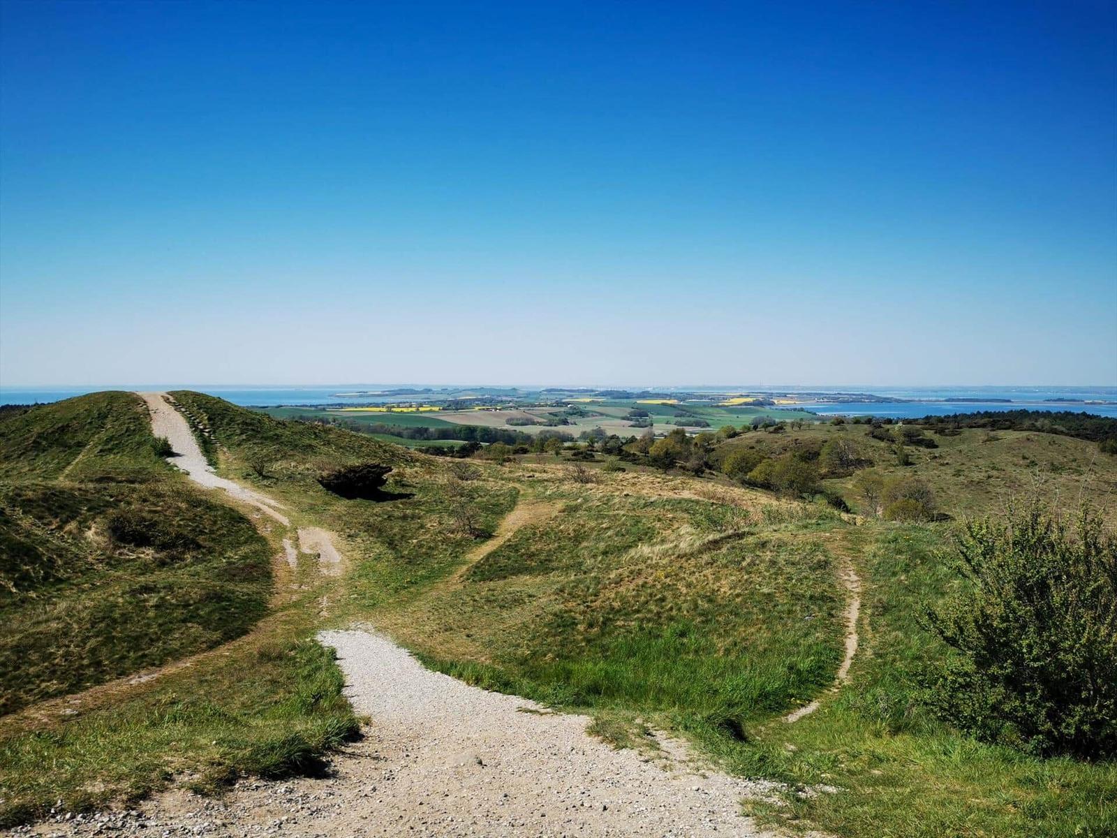 Weg führt über grüne Hügel zu Blick auf Felder und Meer.
