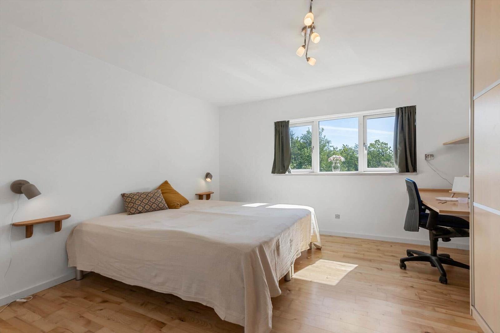 Schlafzimmer mit Bett, Schreibtisch und Fenster mit Blick auf Bäume.
