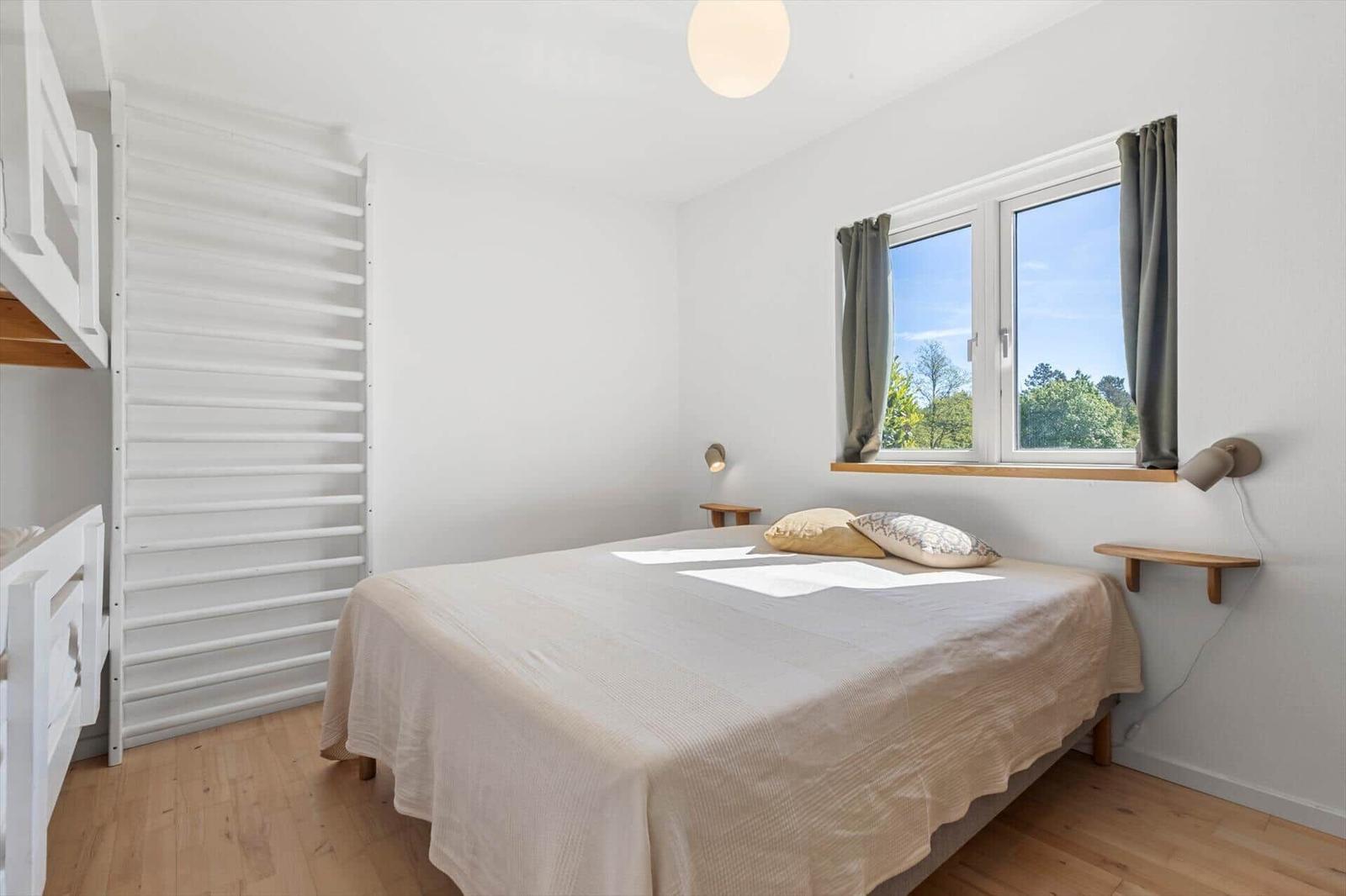 Schlafzimmer mit Bett, Fenster und Holzboden. Links steht ein weißes Hochbett.