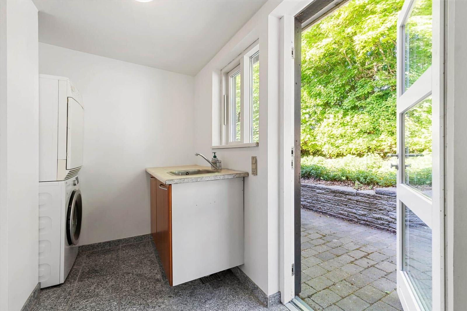 Waschraum mit Waschmaschine, Trockner und Waschbecken. Durchgang zur Terrasse mit Blick auf grüne Bäume.