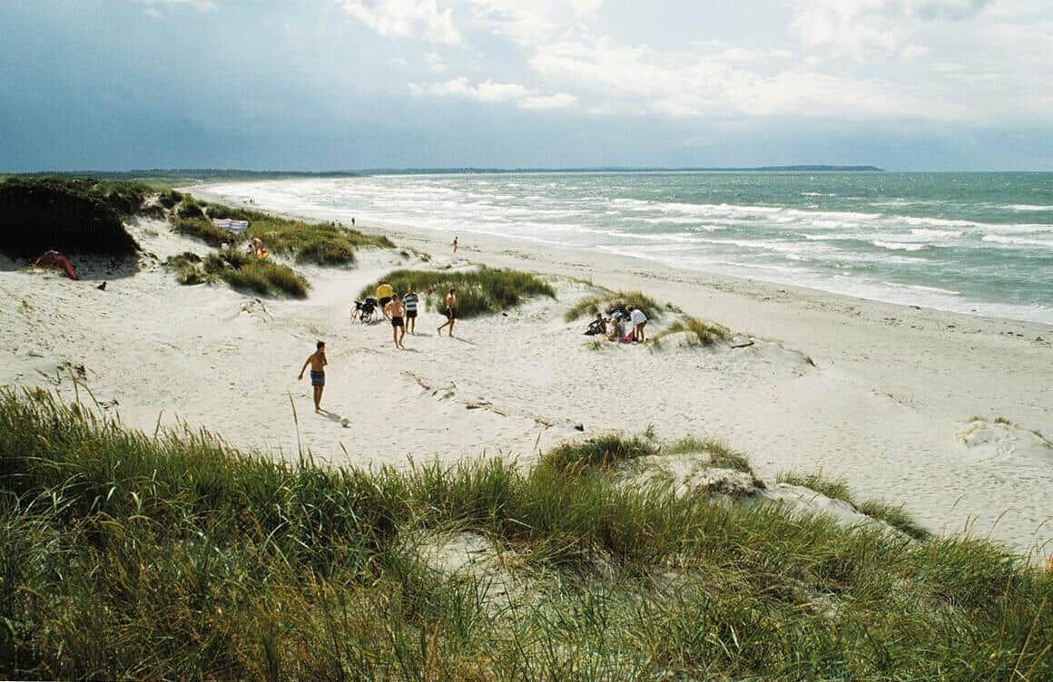 Beach with sand dunes and people walking or sunbathing near the water.