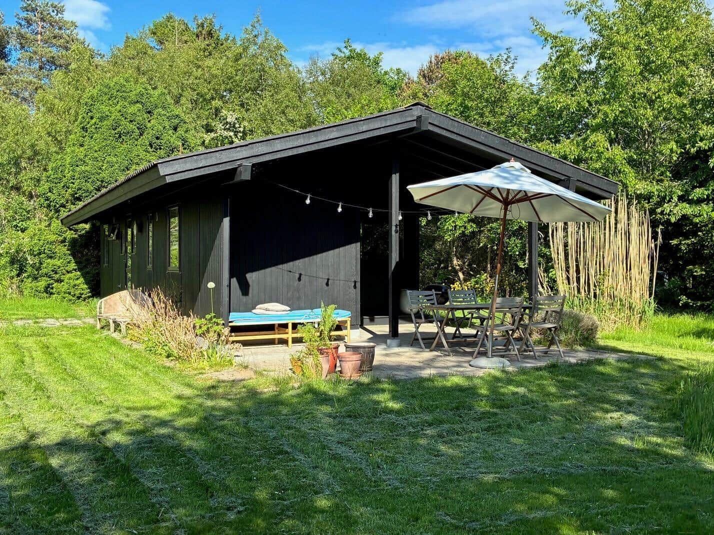 Black wooden house with terrace, sun lounger, and table with chairs under umbrella.