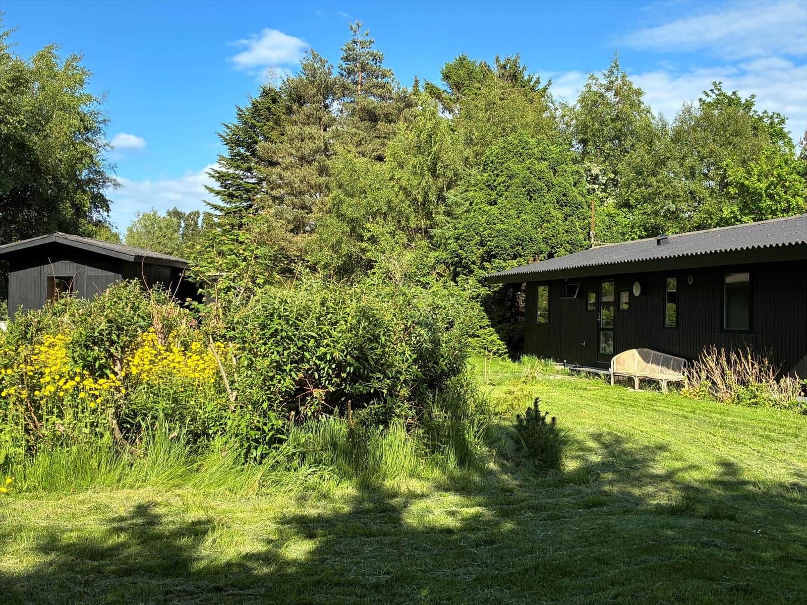 Two dark wooden houses with garden and trees under blue sky.