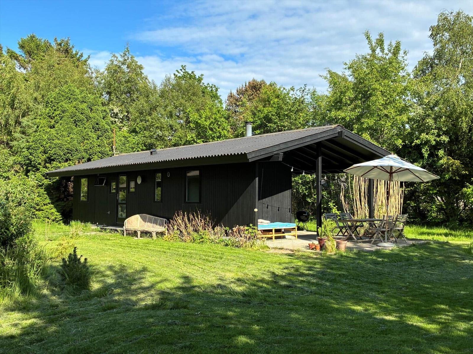 Black cabin with terrace, garden, and umbrella. Surrounded by trees and grass.