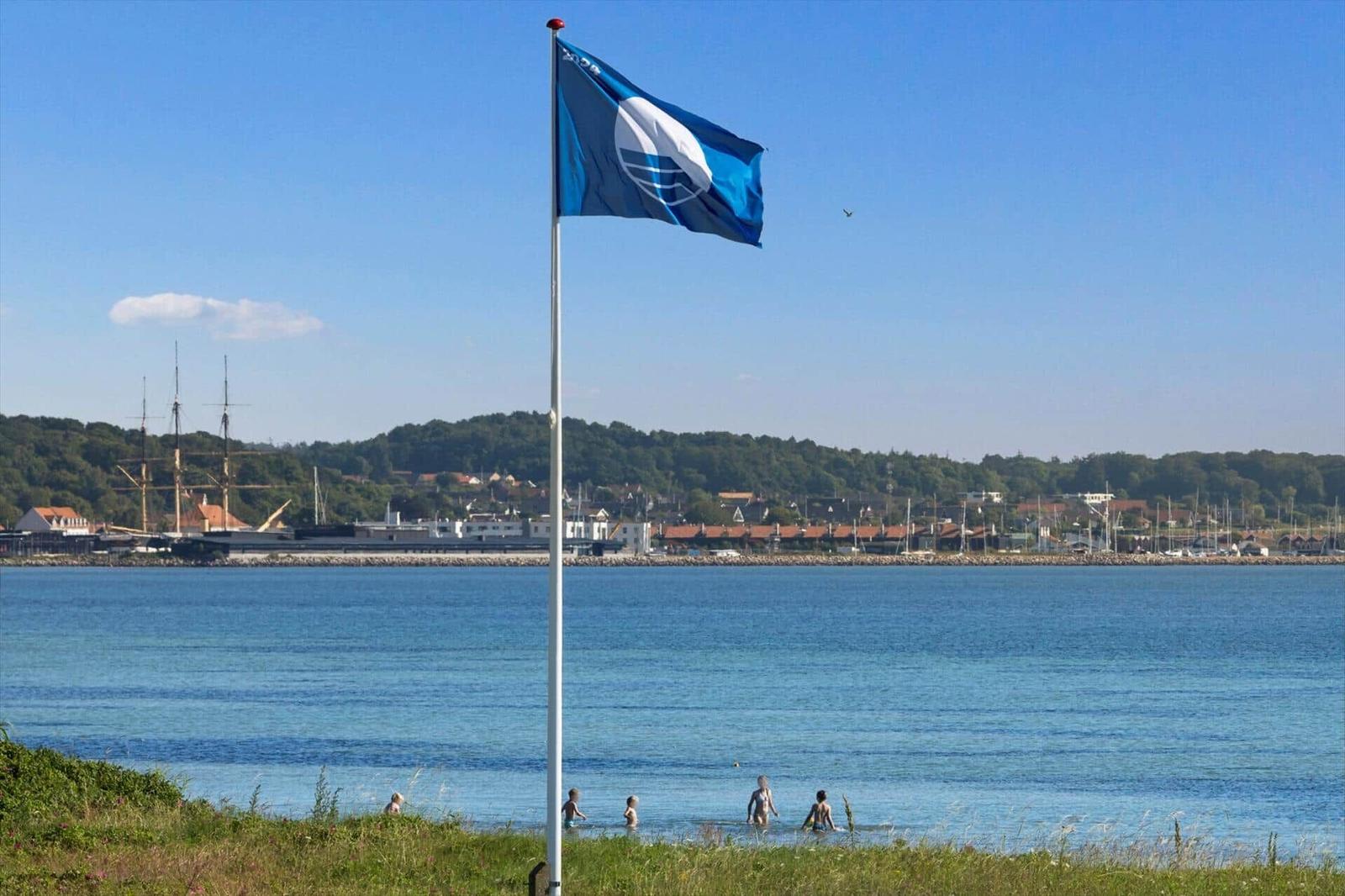 Flag flutters by the shore, people swim in the sea, harbor with ships in the background.