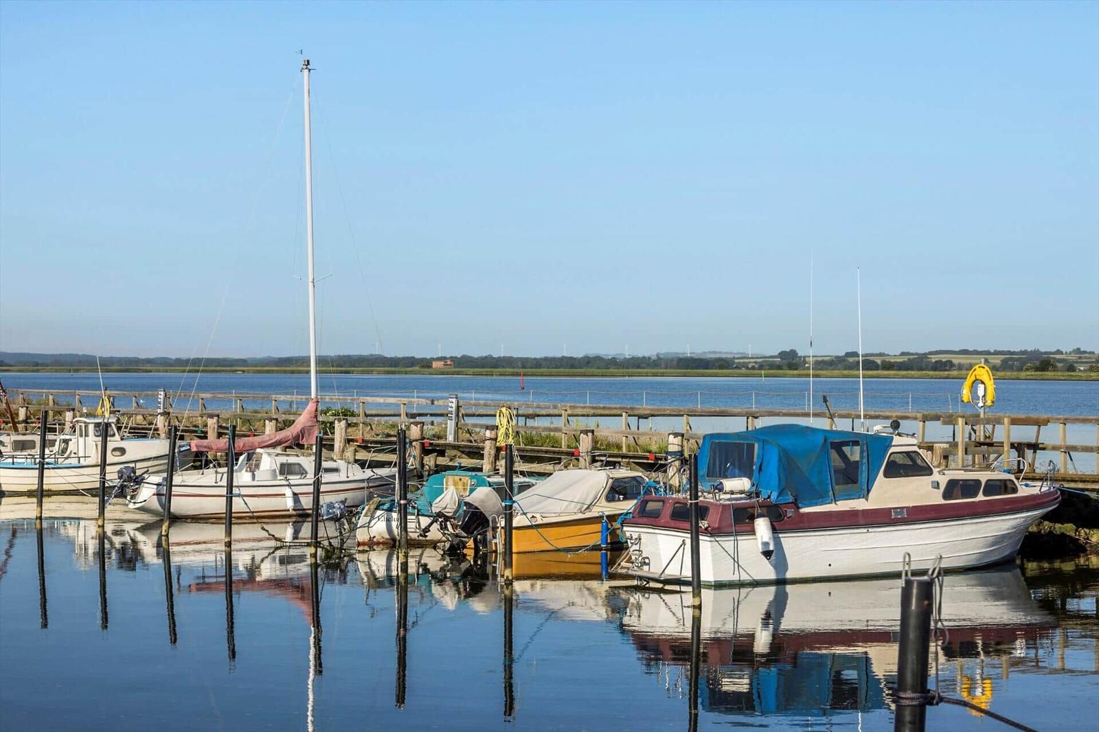 Boats are moored at a wooden pier in the water. In the background, fields and a clear sky are visible.