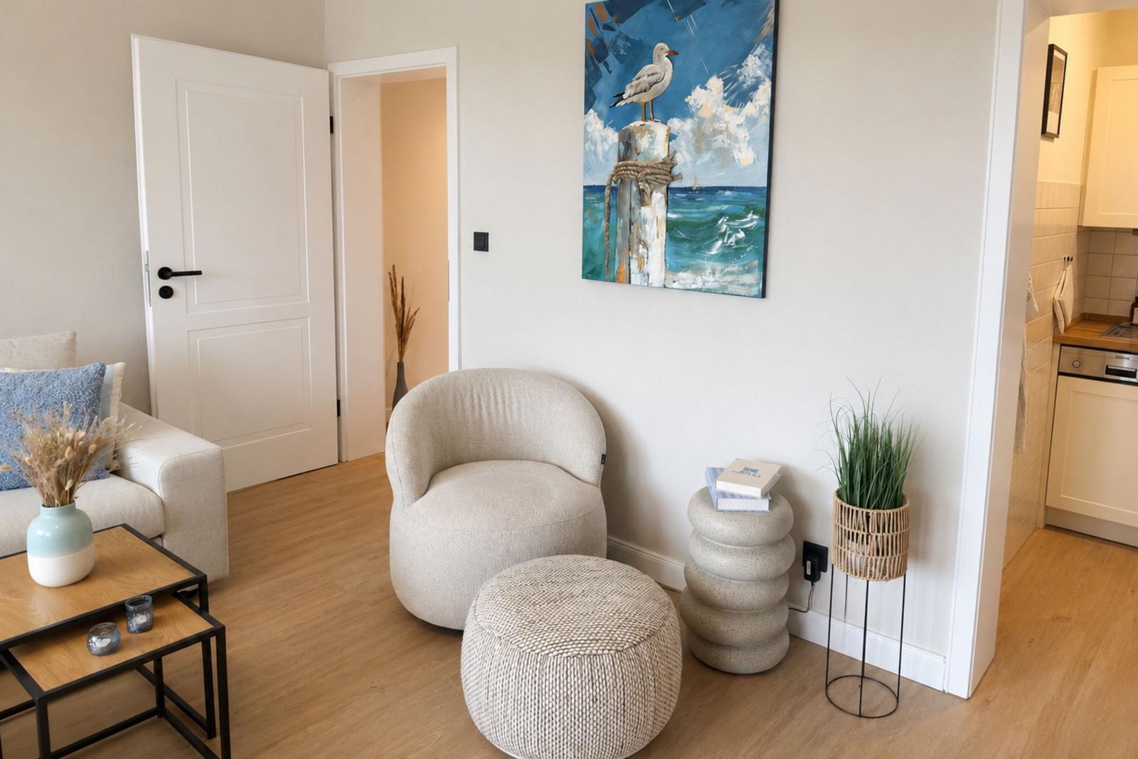 Wohnzimmer mit Sitzgelegenheit, Wandbild und Pflanze. Küchenbereich sichtbar.
