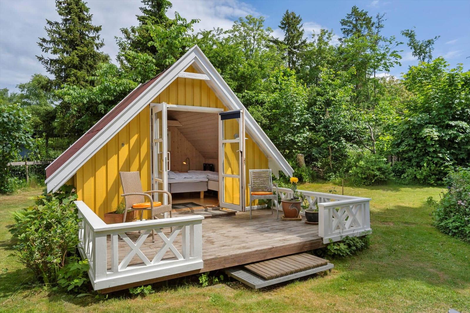 Gelbes Holzhaus mit offener Tür, Balkon und Bett im Inneren, umgeben von Grün.