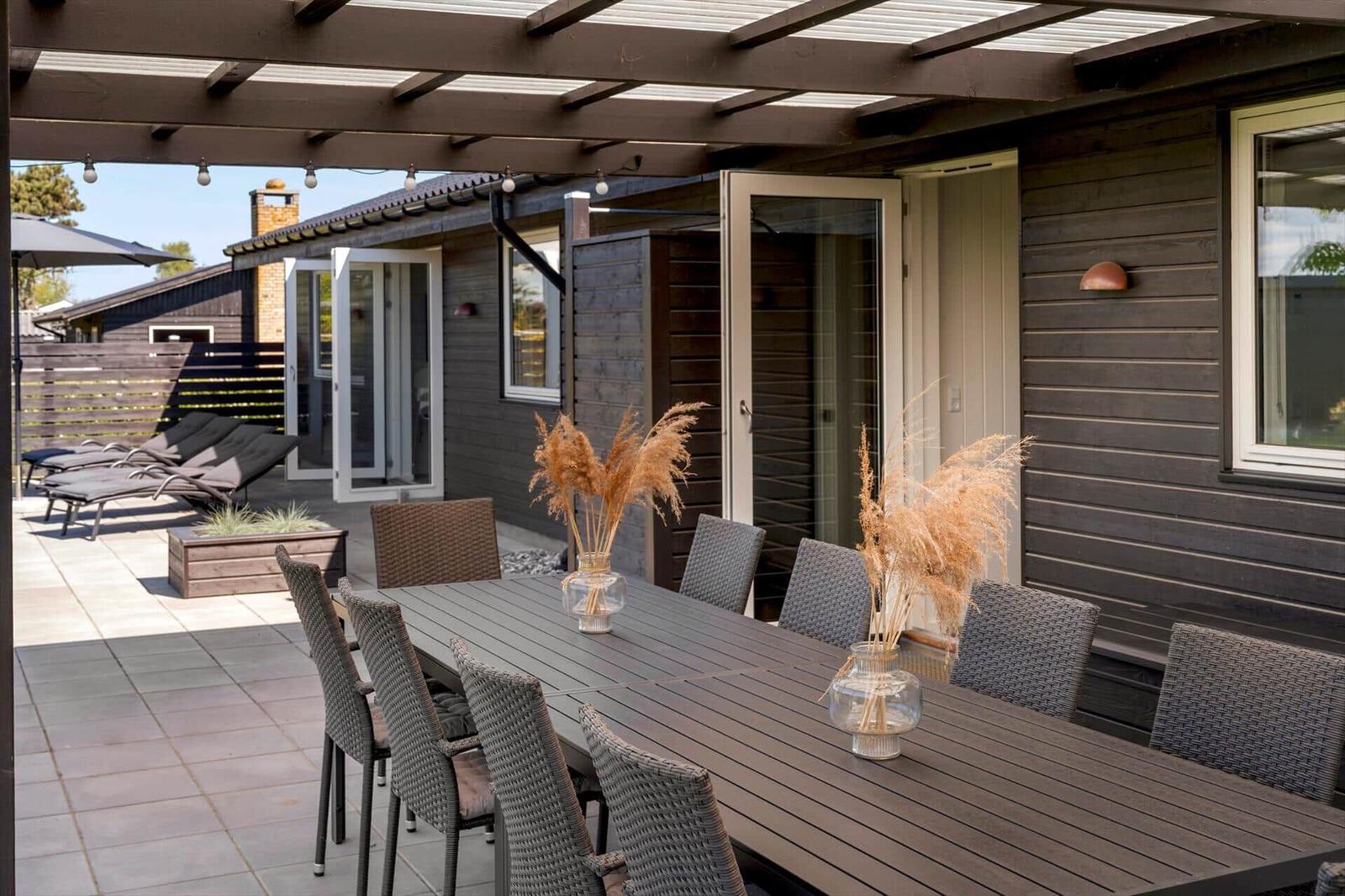 Terrasse mit Esstisch, Stühlen und Sonnenschirm. Holzwände und Glastüren führen ins Haus.