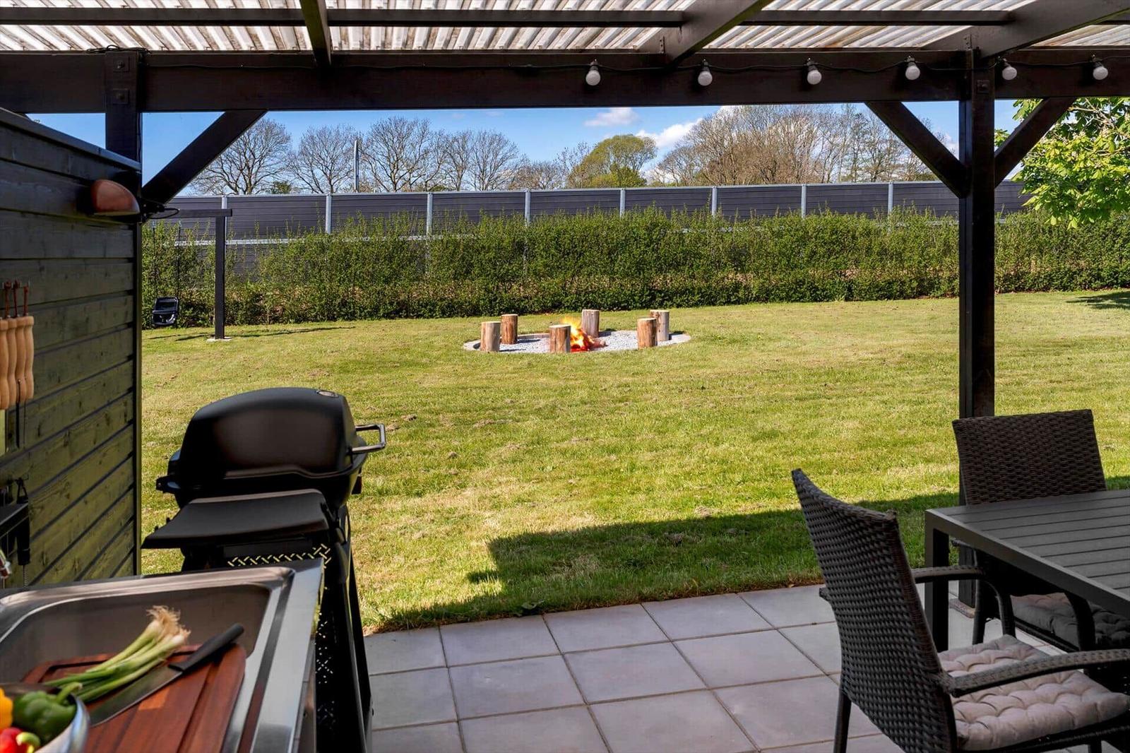 Terrasse mit Grill, Tisch und Stühlen. Im Hintergrund brennt ein Feuer im Garten.