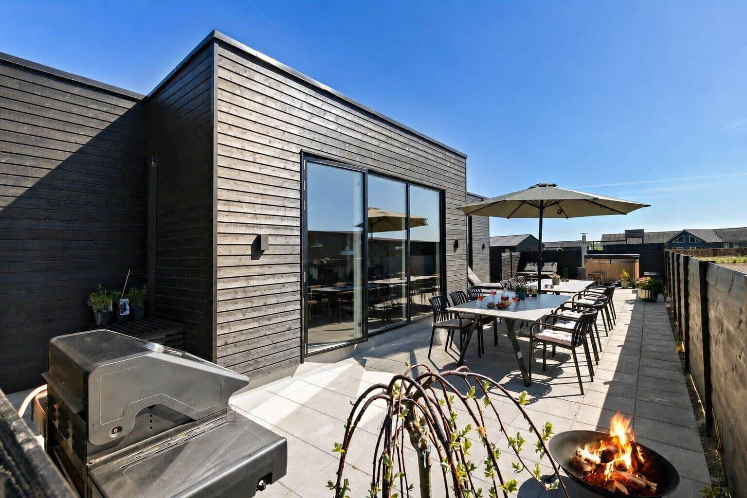 Terrasse mit Grill, Tisch, Stühlen und brennendem Feuerkorb. Modernes Haus mit Glasfront.