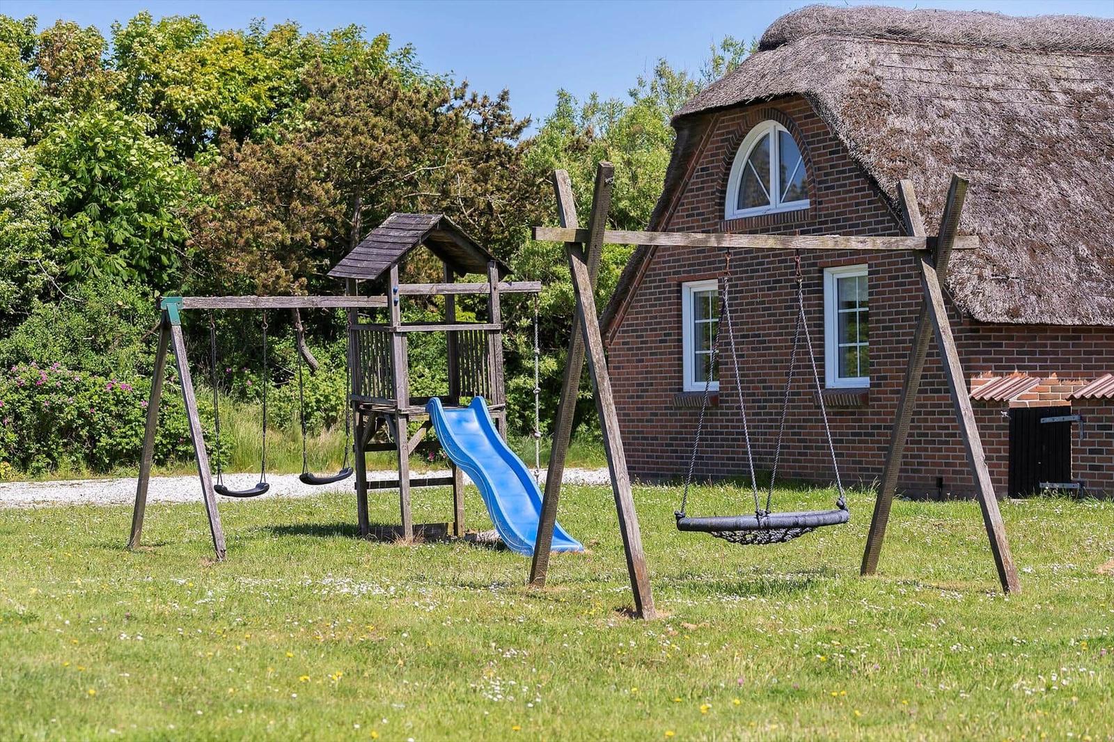 Ein Spielplatz mit Schaukel und Rutsche vor einem strohgedeckten Haus.