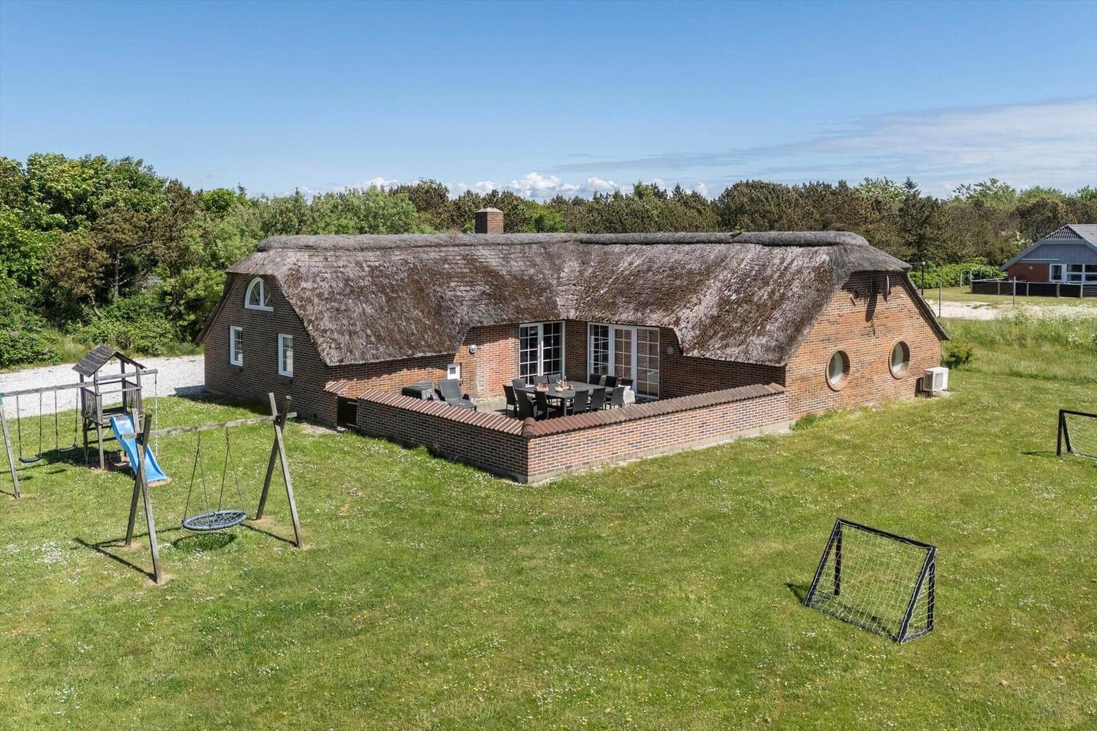 Haus mit Strohdach, Terrasse, Spielplatz und Fußballtor auf grünem Grund.
