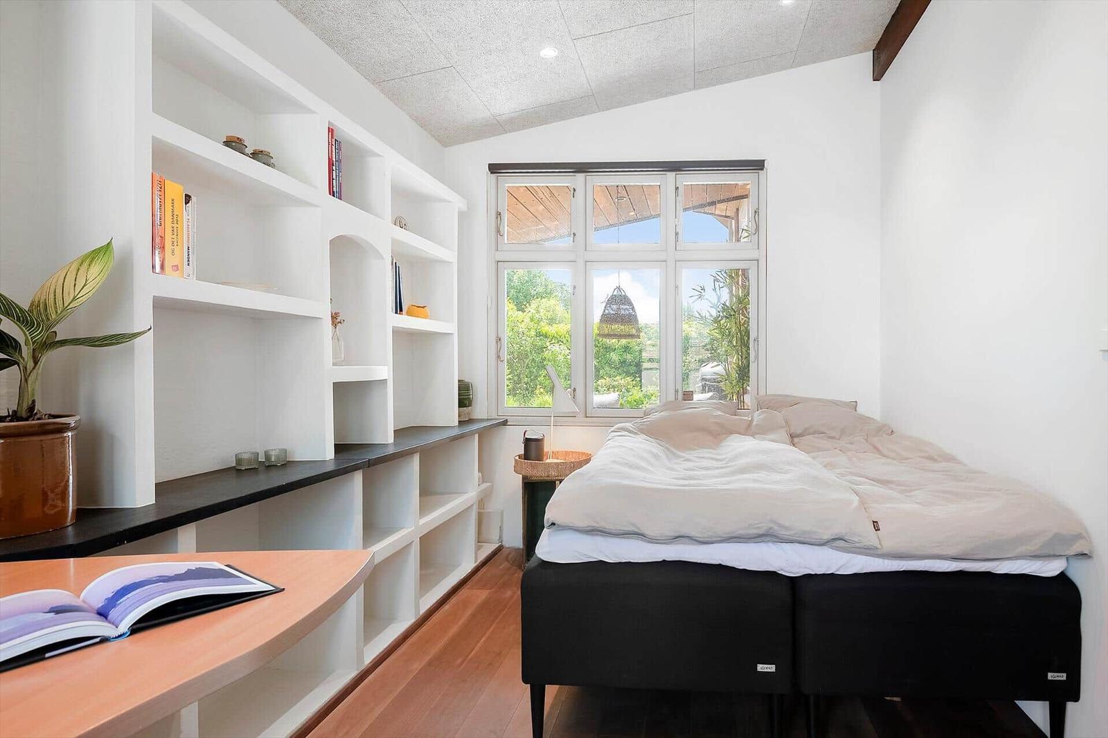 Bedroom with bed, window, and bookshelf. Wooden floor and plant visible.