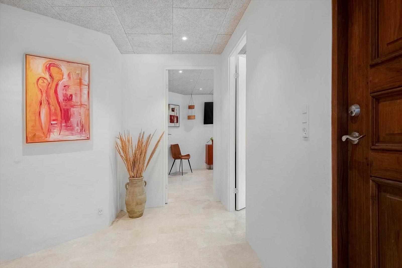 Hallway with wall art, vase, and view into living area with chair and TV.
