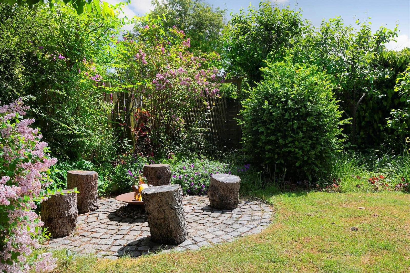 Garden with fire pit and log seats on stone pavers, surrounded by green shrubs and lilac flowers.