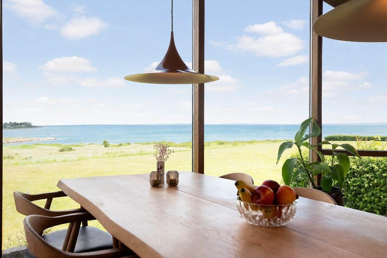 Dining room with wooden table, fruit bowl, and view of sea and green field.