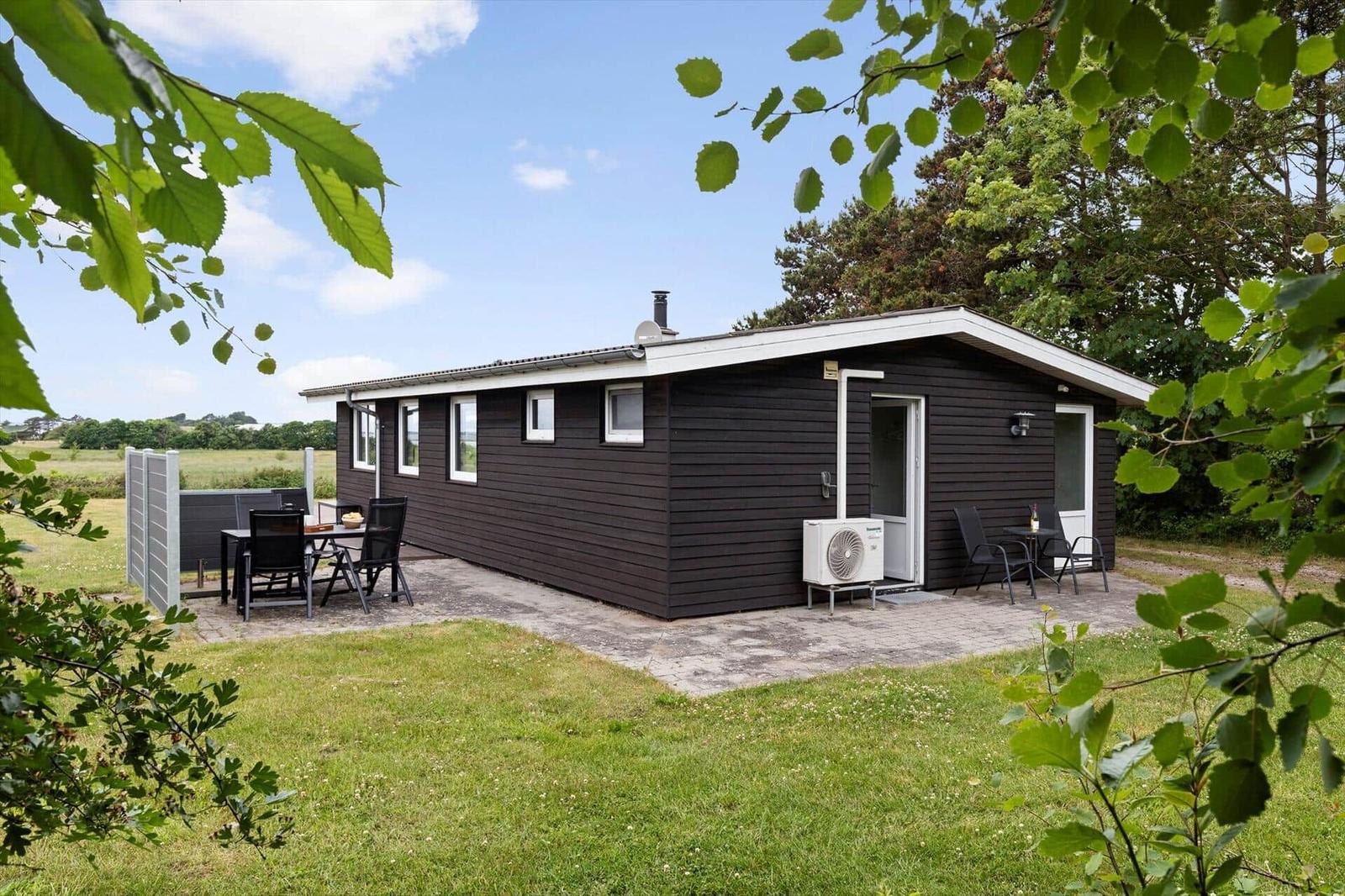 Black wooden house with terrace, garden, and seating. Background: green fields and trees.