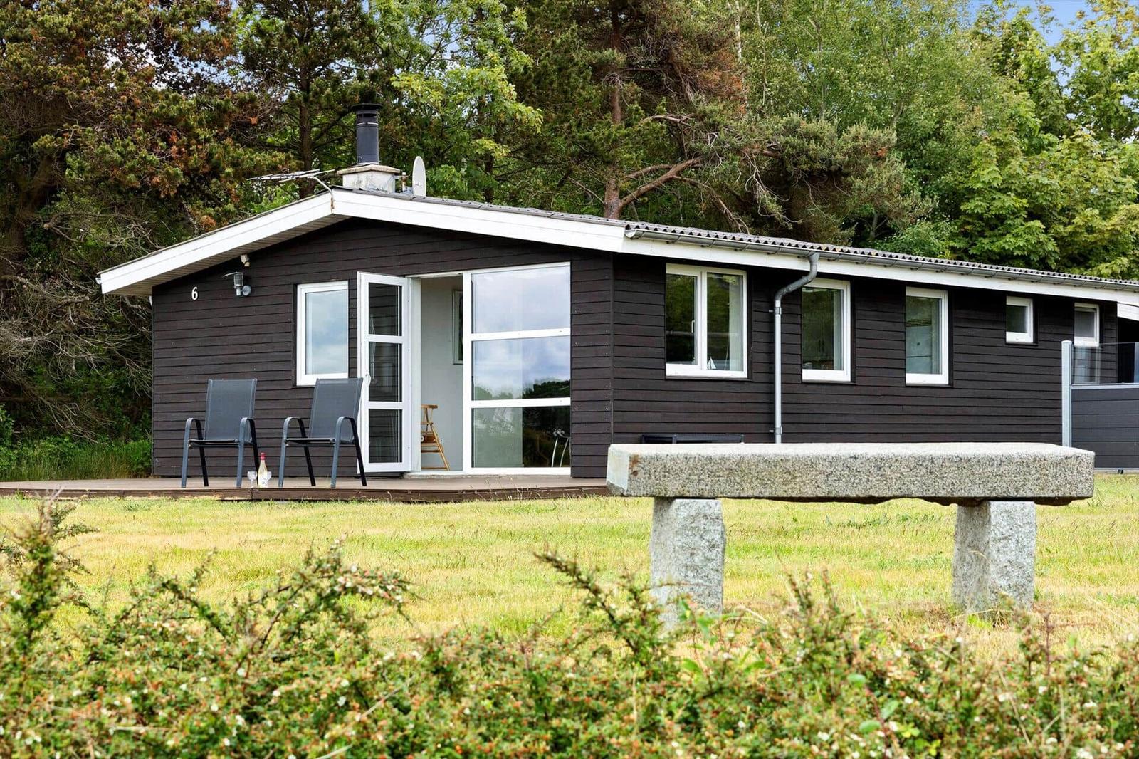 A dark brown holiday home with white trim and a terrace. A stone bench is in the foreground.