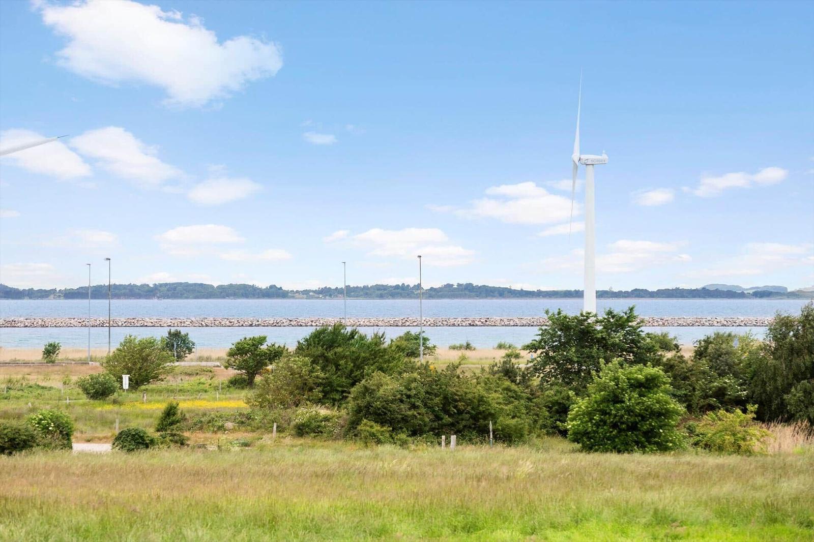Wind turbine by water with green areas and trees in foreground.