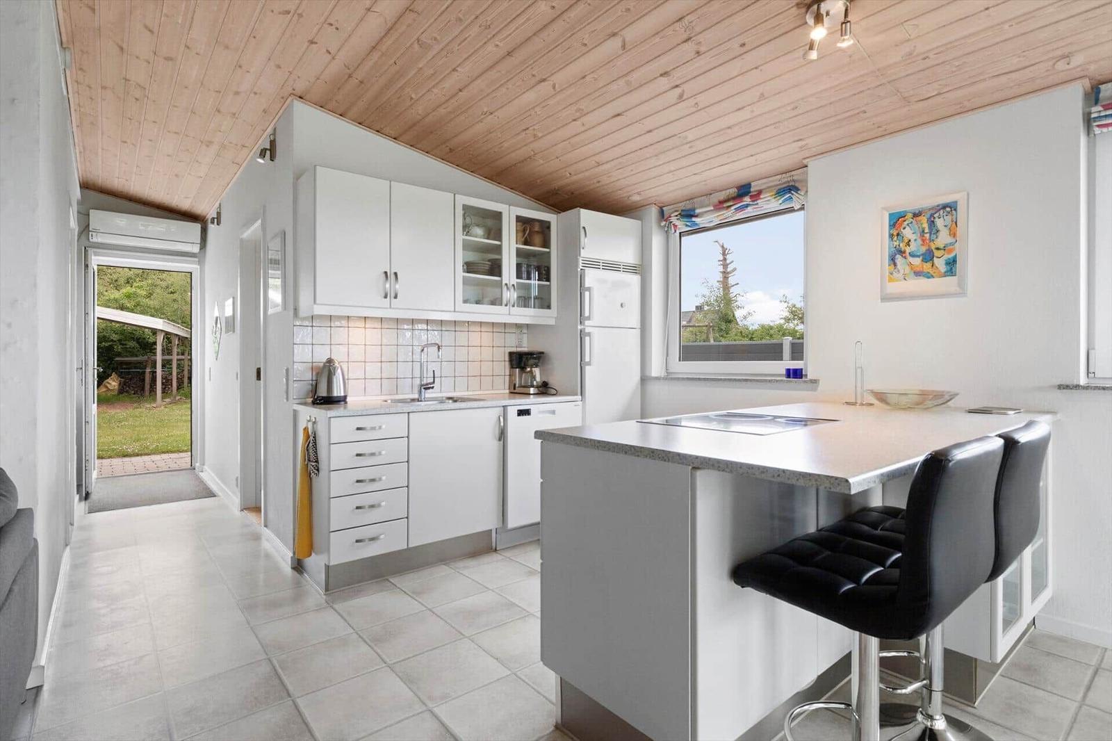 Kitchen with white cabinets, kitchen island, and black bar stools. Wooden ceiling and window with view outside.