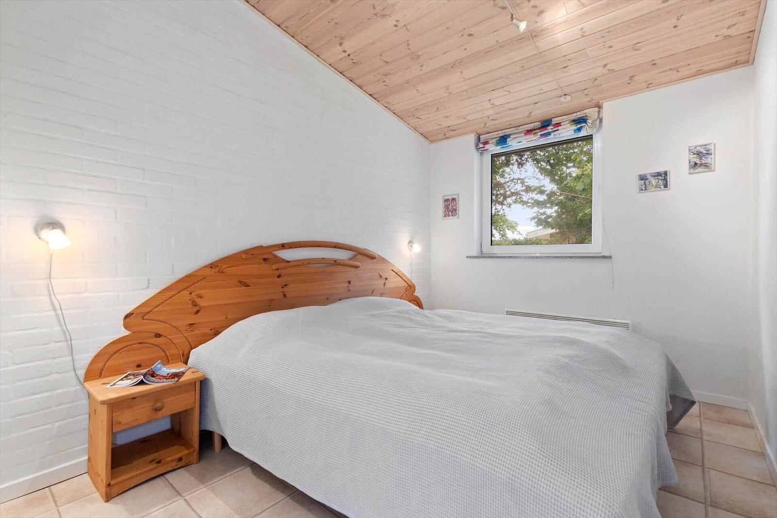 Bedroom with wooden bed, white wall, and wooden ceiling. Window with view outside.