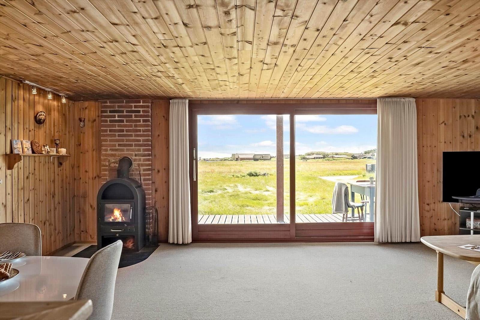 Wohnraum mit Holzdecke, Kamin und großem Fensterblick auf die Wiese.