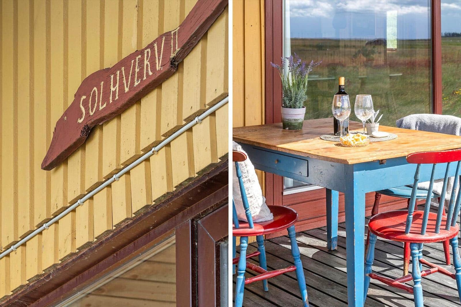Holztafel mit Schriftzug SOLHVERV II an gelbem Holzhaus. Terrasse mit Tisch, Stühlen und Blick auf Feld.