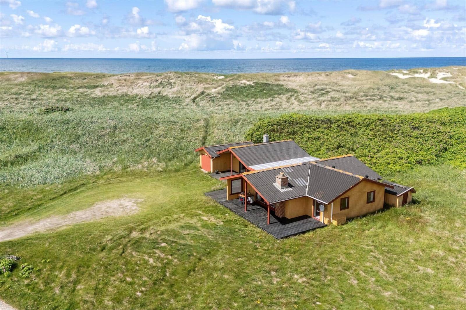Holzhäuschen mit Terrasse, umgeben von Gras und Dünen, mit Blick auf das Meer.