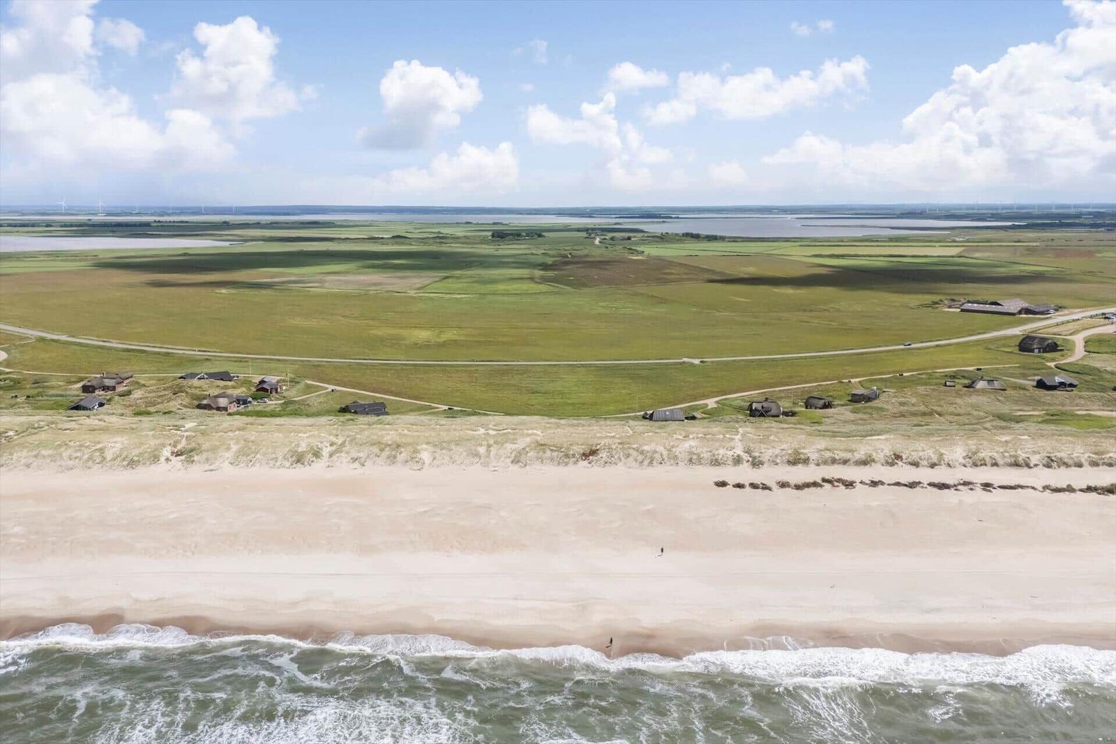 Strand mit Dünen, grünen Feldern und wenigen Häusern unter blauem Himmel.