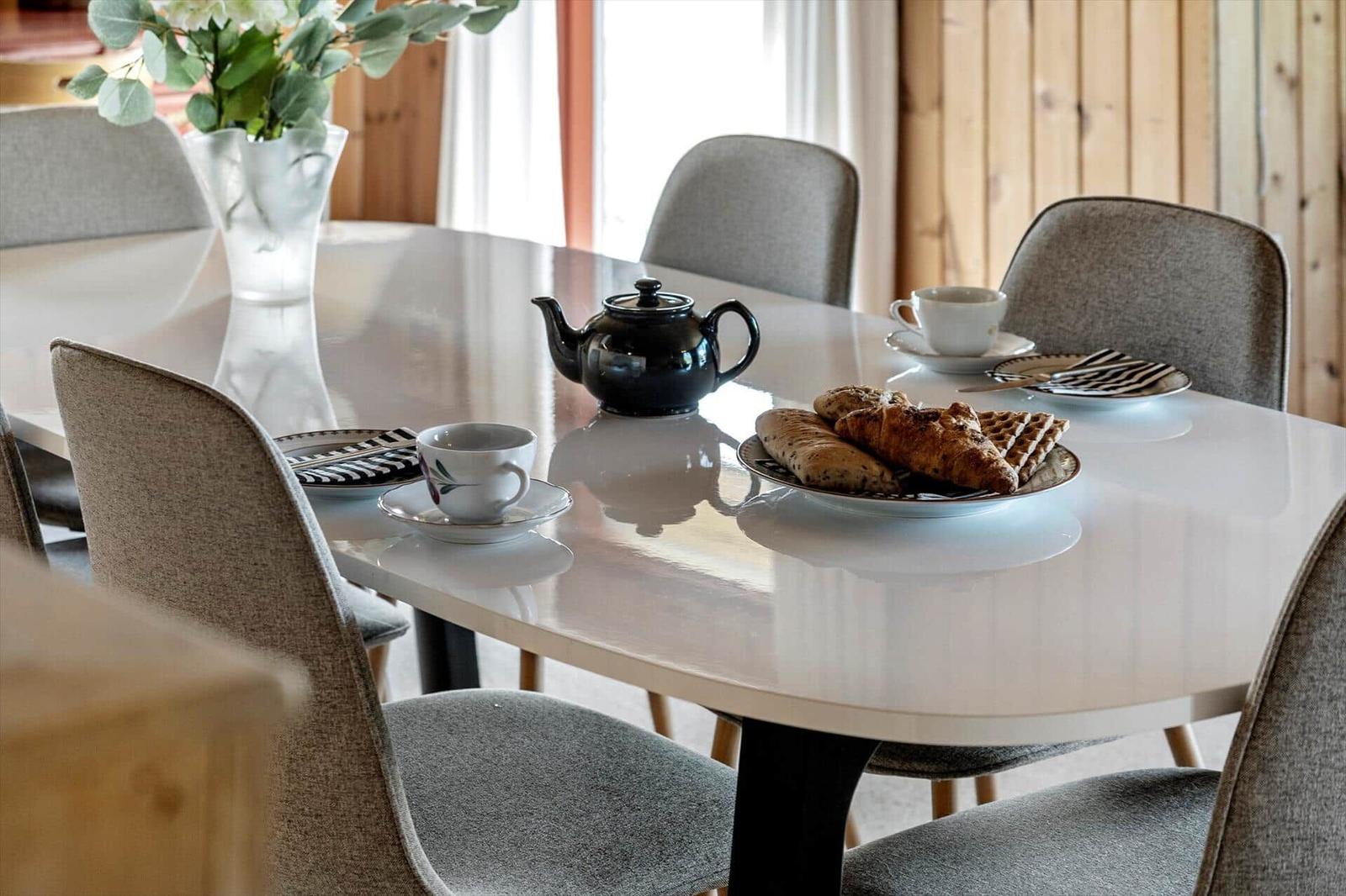 Runde Tisch mit Stühlen, Teekanne, Tassen und Brötchen. Holzwand im Hintergrund.