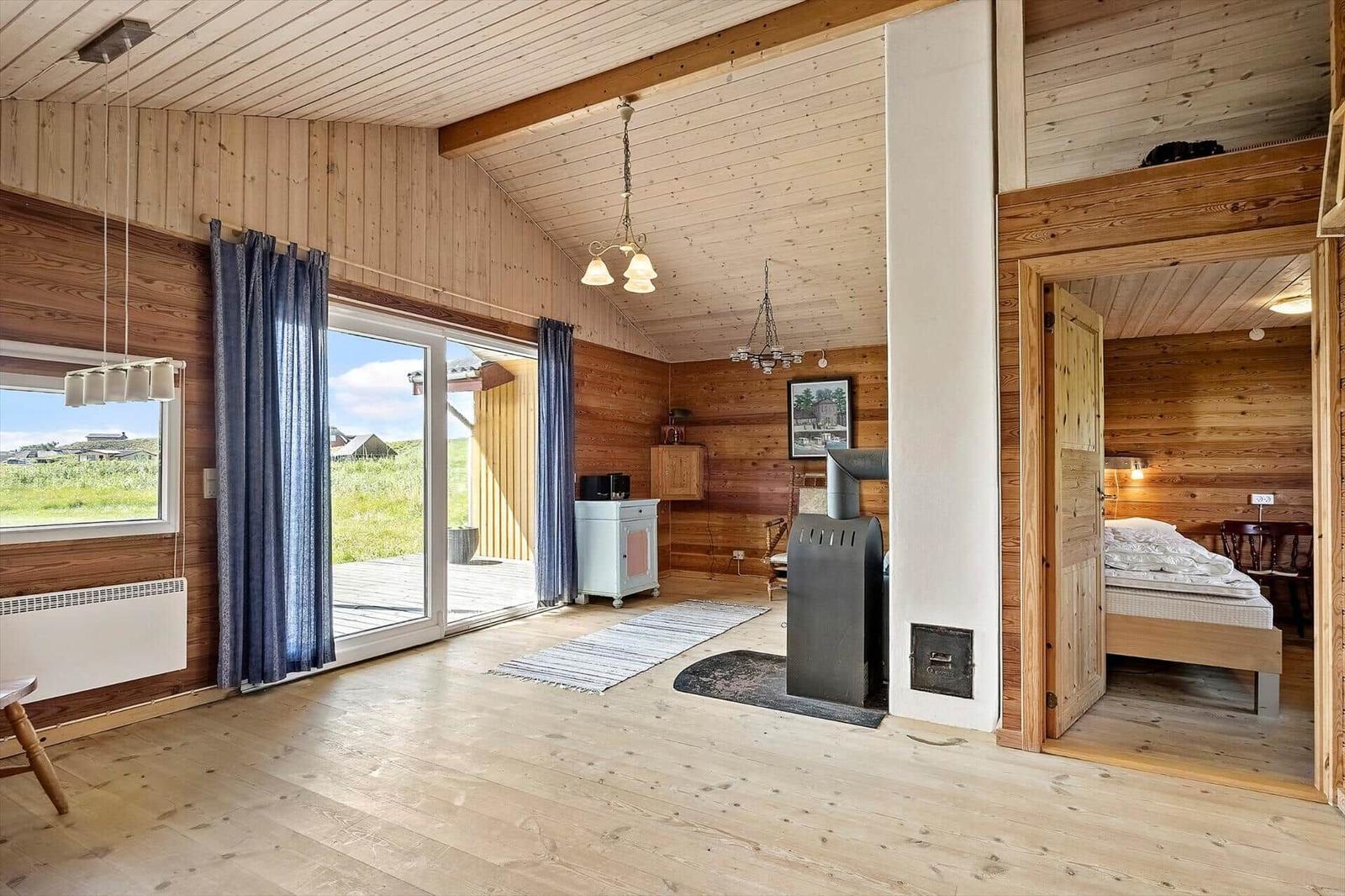 Wohnraum mit Holzverkleidung, Kamin und Blick auf Terrasse und Grünland.