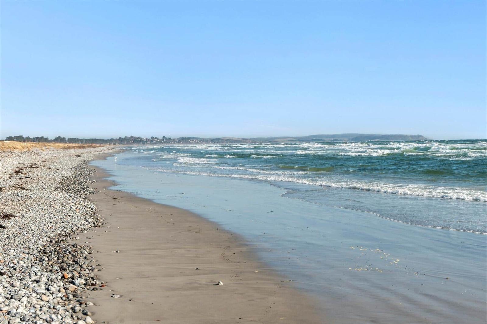 Strand mit Kies und Sand, Wellen brechen am Ufer, Horizont mit Hügeln im Hintergrund.