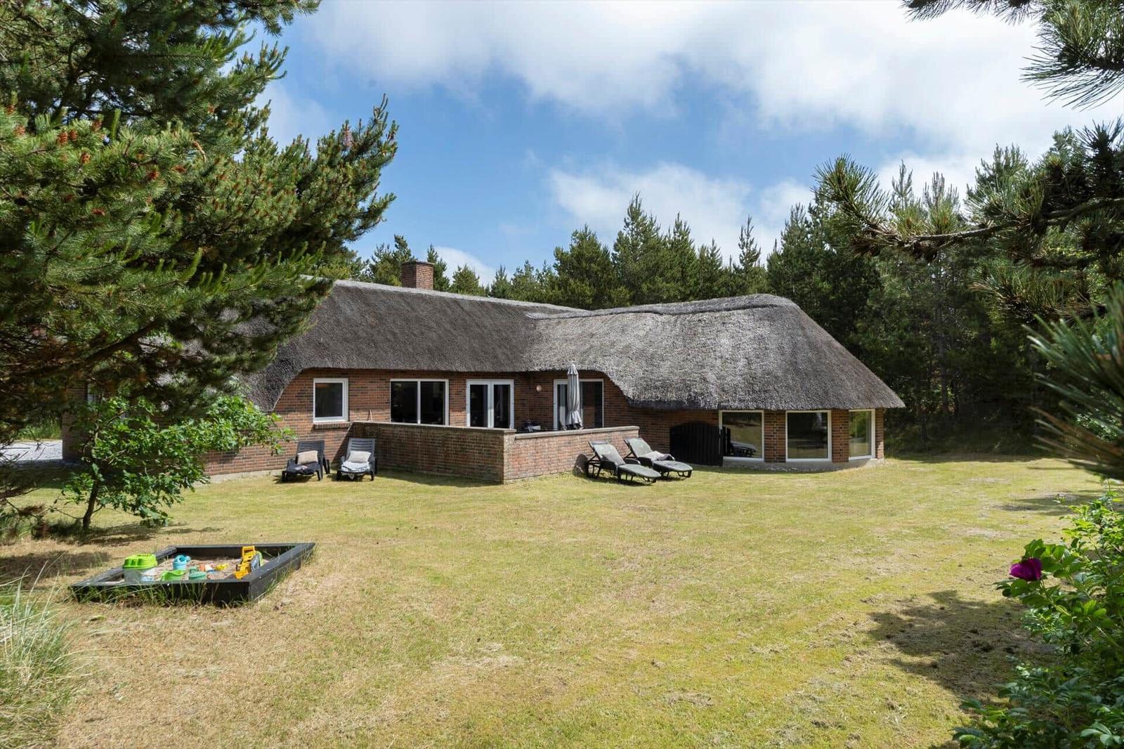 Haus mit Strohdach, Garten, Liegestühle und Spielplatz. Umgeben von Nadelbäumen.