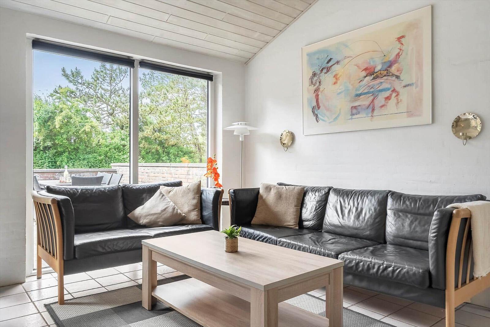 Zwei schwarze Ledersofas stehen um einen Holztisch herum. Große Fenster zeigen einen grünen Außenbereich.