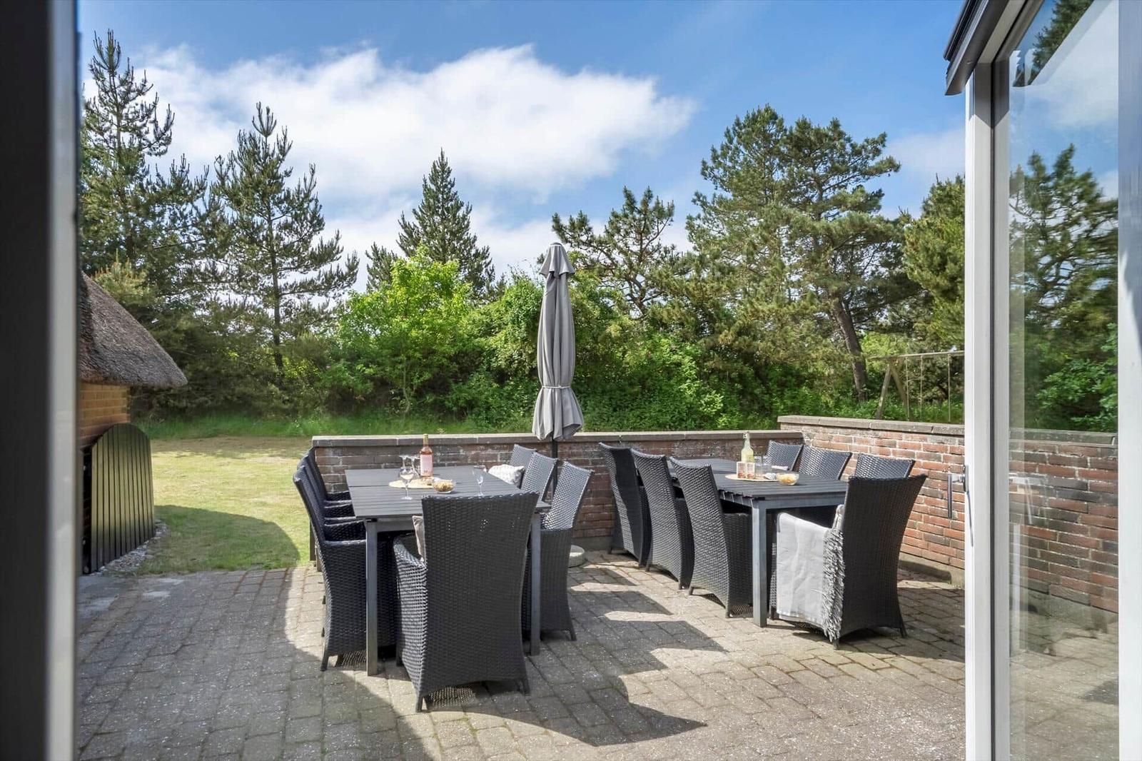 Terrasse mit zwei Tischen, Stühlen und einem geschlossenen Sonnenschirm. Hintergrund: Bäume und Himmel.