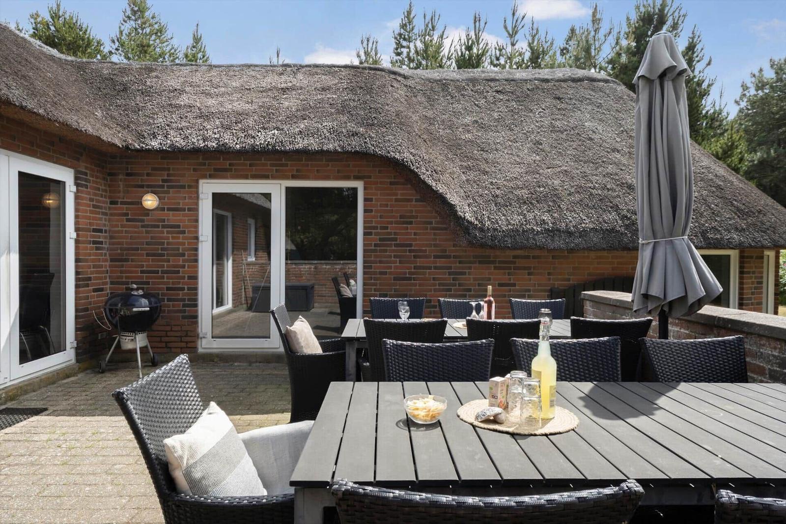 Terrasse mit Tisch, Stühlen und Sonnenschirm vor einem Haus mit Strohdach und Backsteinwand.