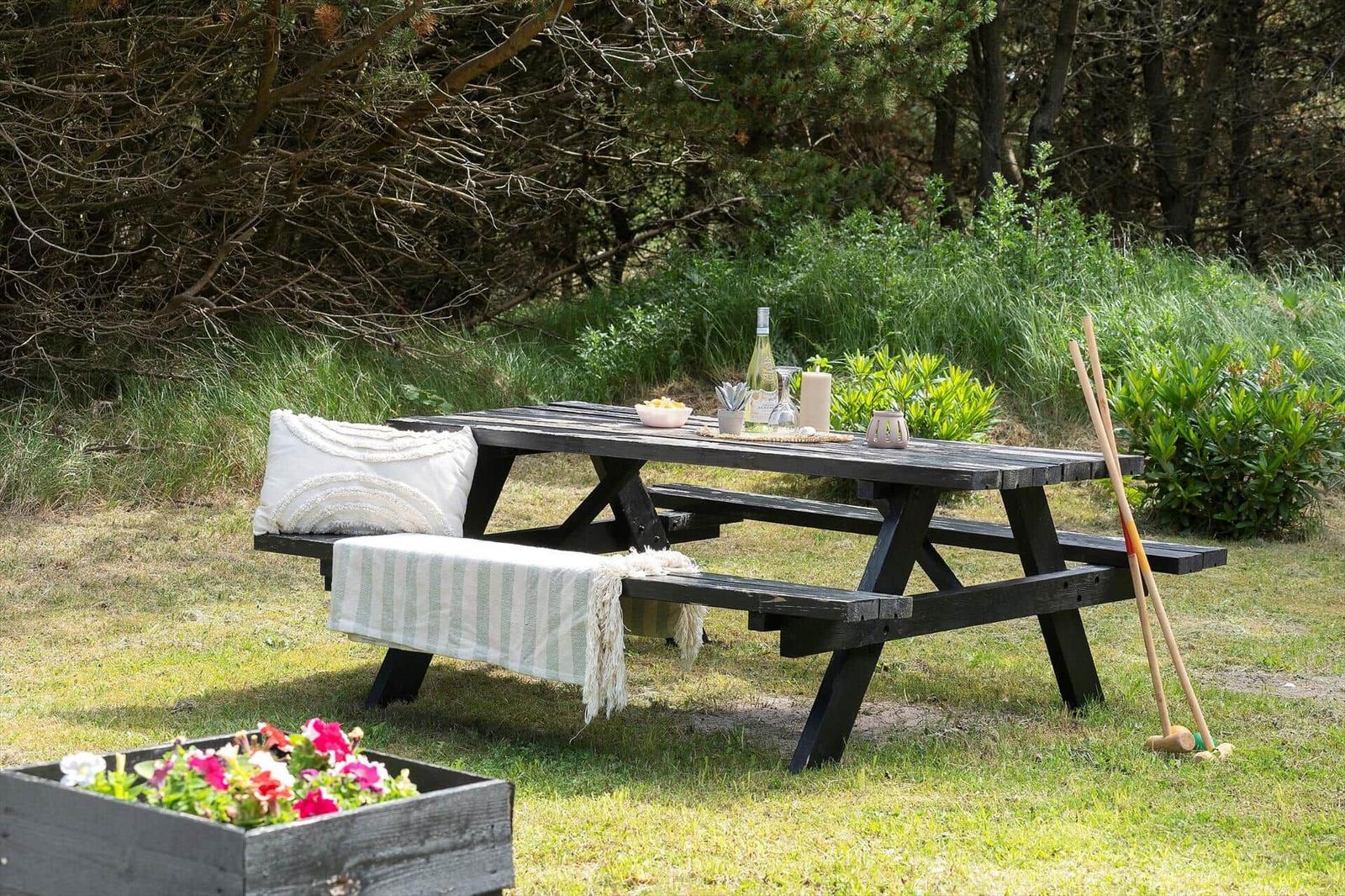 Holzpicknicktisch mit Decke und Kissen im Garten. Hintergrund: Gras und Bäume.