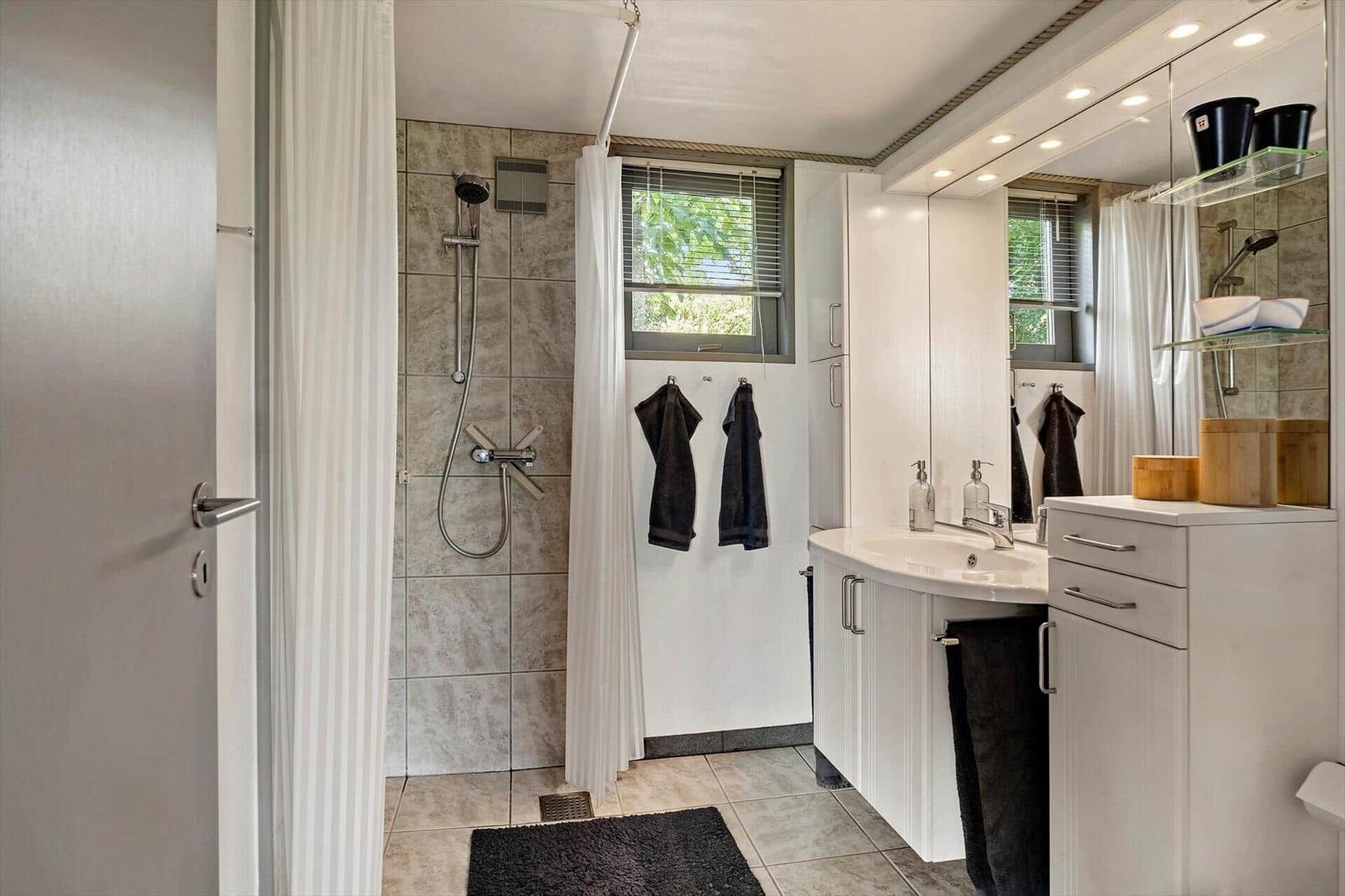 Bathroom with shower, sink, and mirror cabinet. Window with blinds and towels.