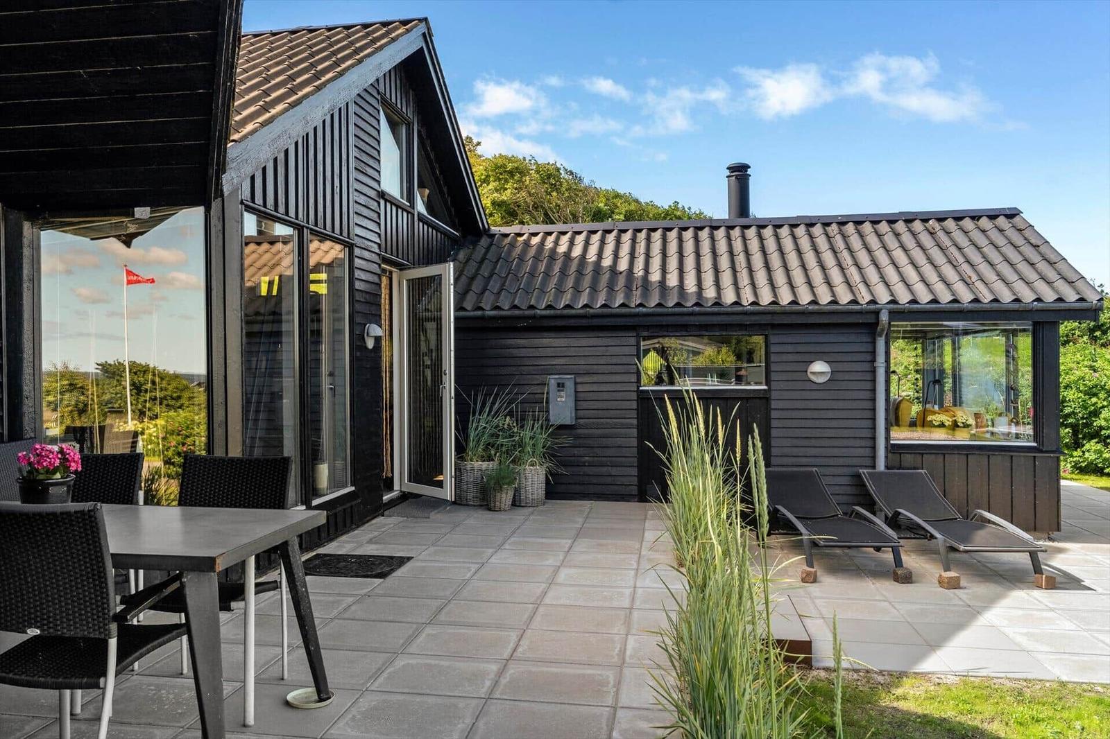 Black wooden house with terrace, sun loungers, and garden. Large windows and roof overhang.