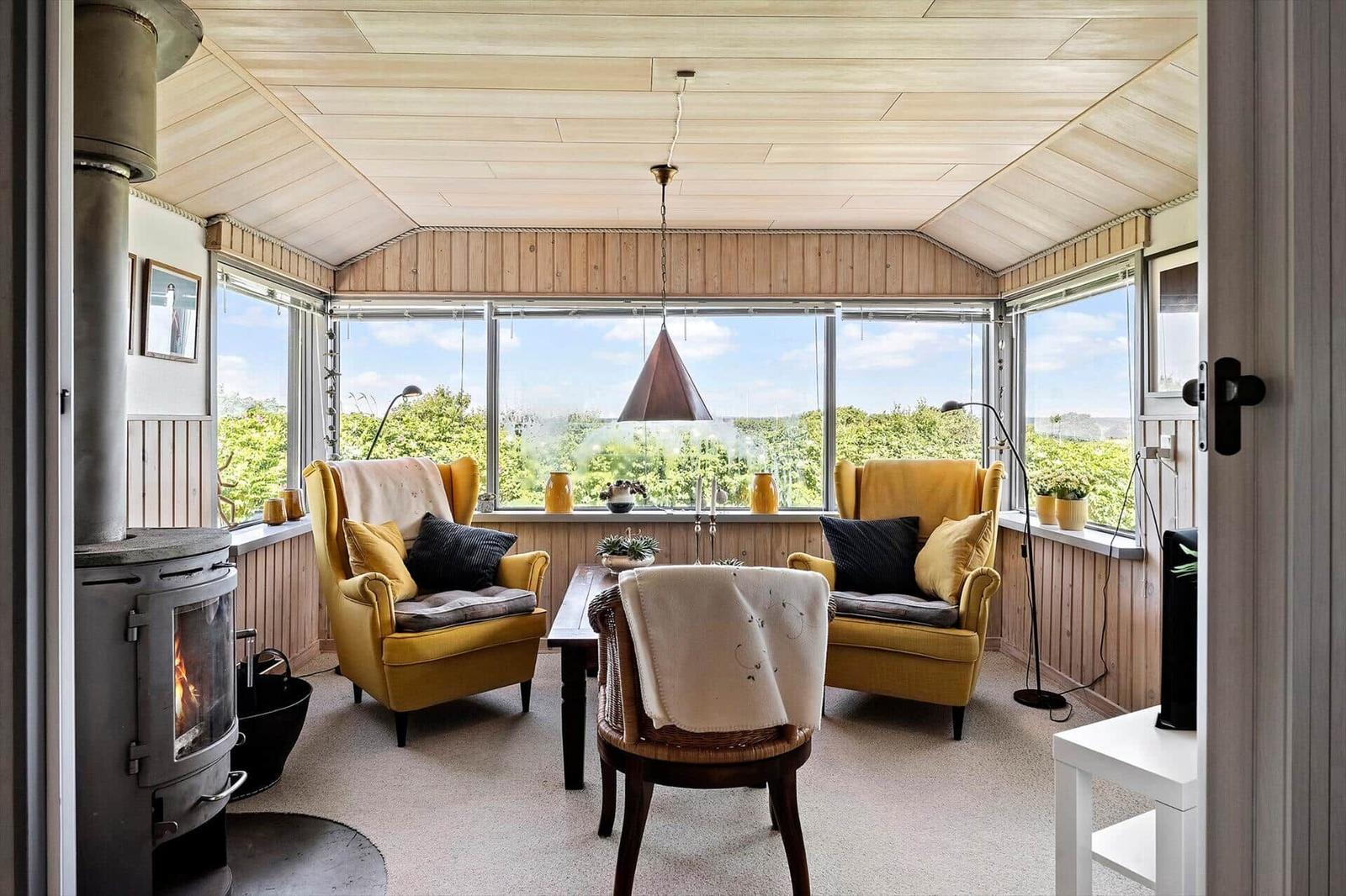 A room with two yellow armchairs, a stove, and large windows overlooking greenery.