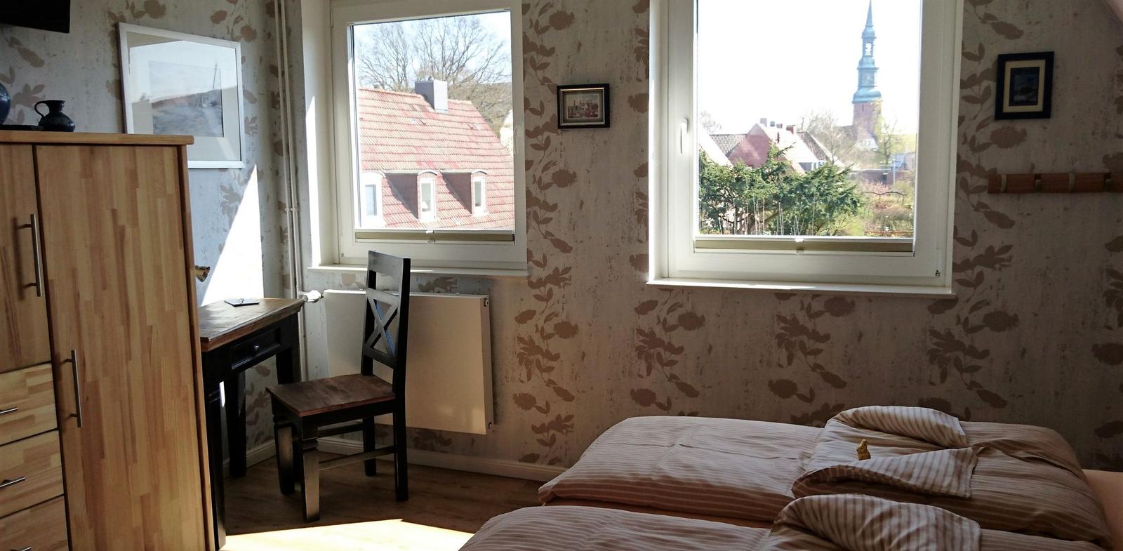 Schlafzimmer mit Bett, Schreibtisch und Fenstern zum Blick auf Dächer und Turm.